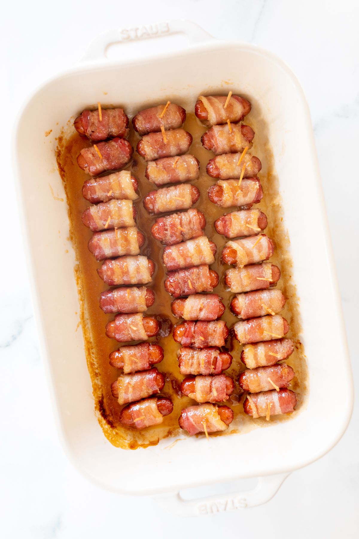 A white baking dish lined with bacon wrapped brown sugar little smokies.
