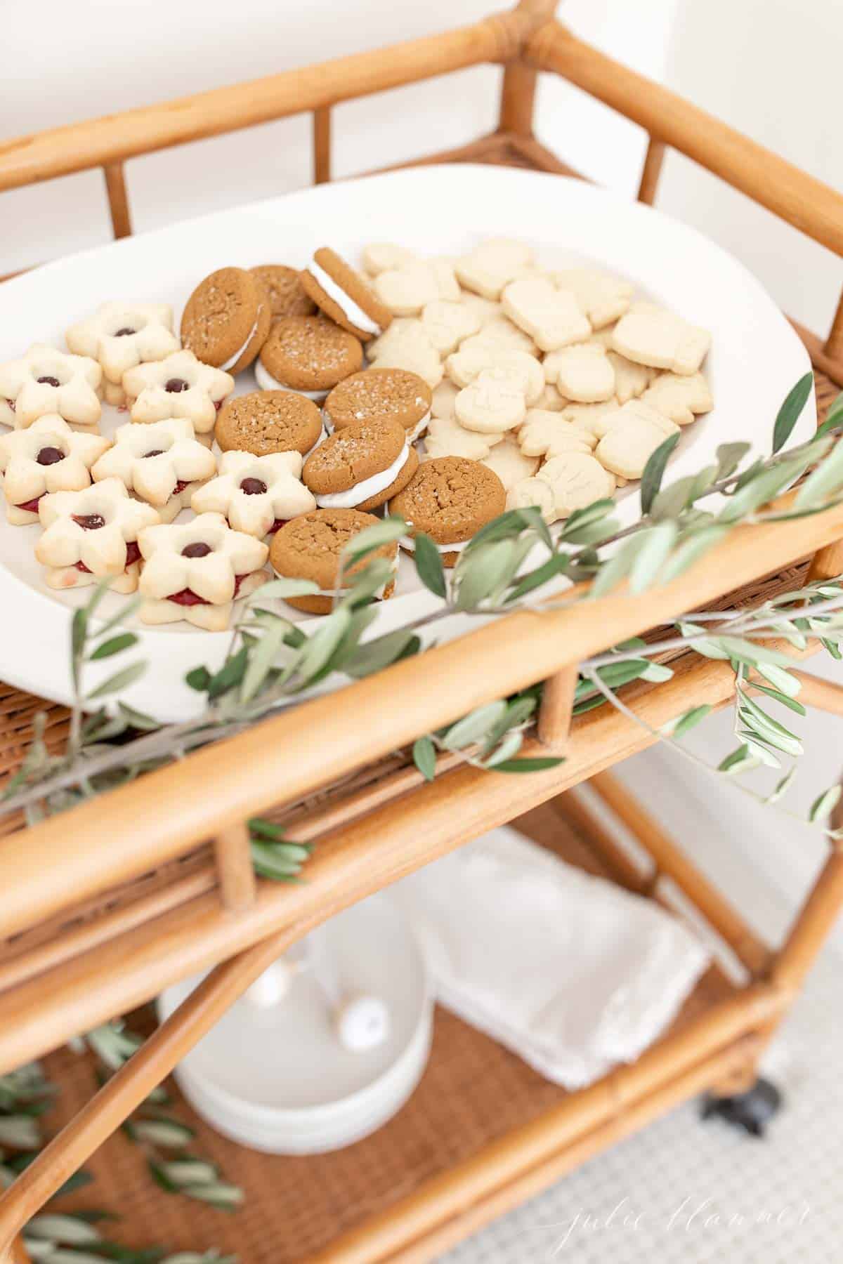 A bamboo bar cart with a white oval platter filled with Scandinavian Christmas cookies, fresh greenery accents.