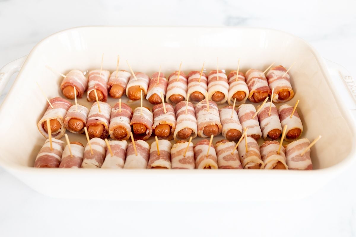 A white baking dish lined with bacon wrapped brown sugar little smokies before baking.