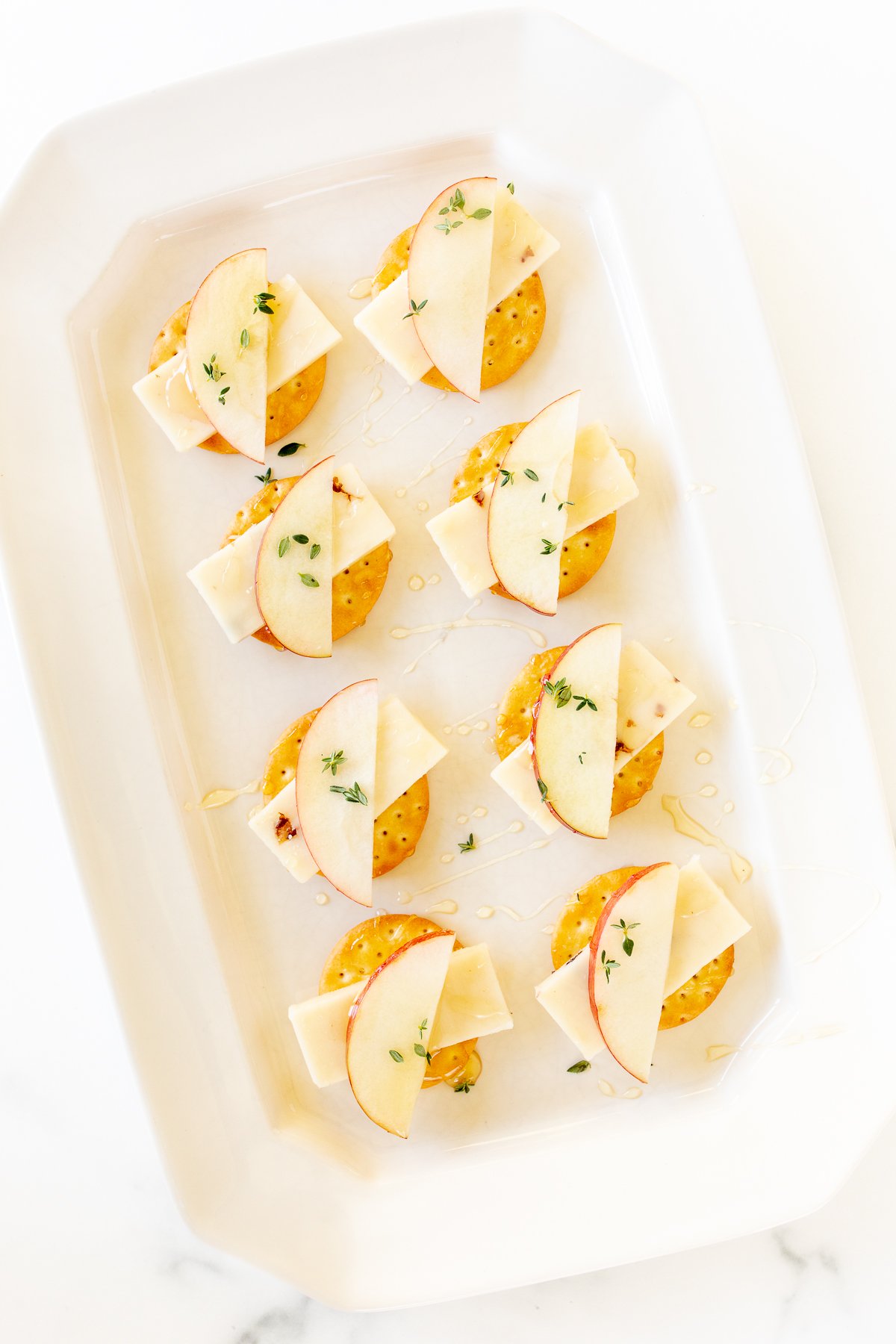 Cheese and crackers on a white platter, with an apple slice and fresh herbs. 