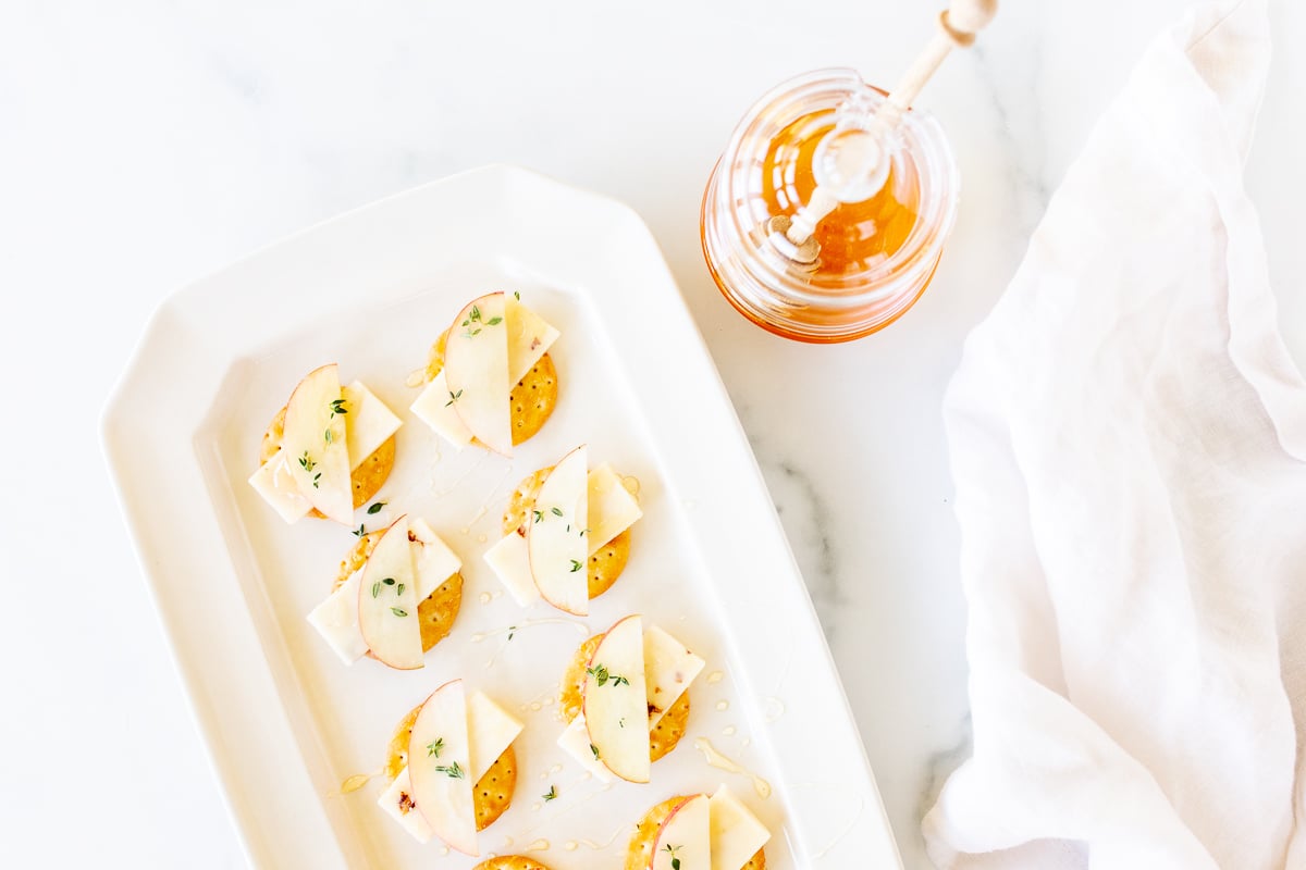 Cheese and crackers on a white platter, with an apple slice and fresh herbs.