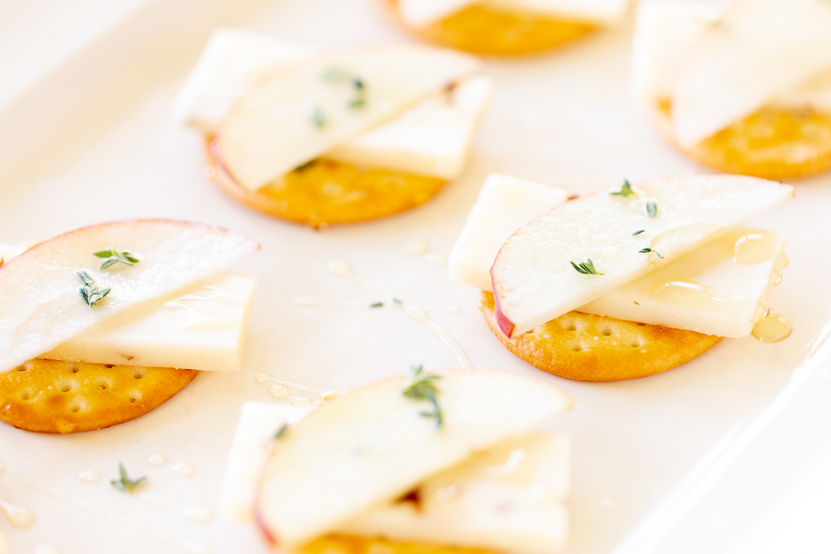 Cheese and crackers on a white platter, with an apple slice and fresh herbs.