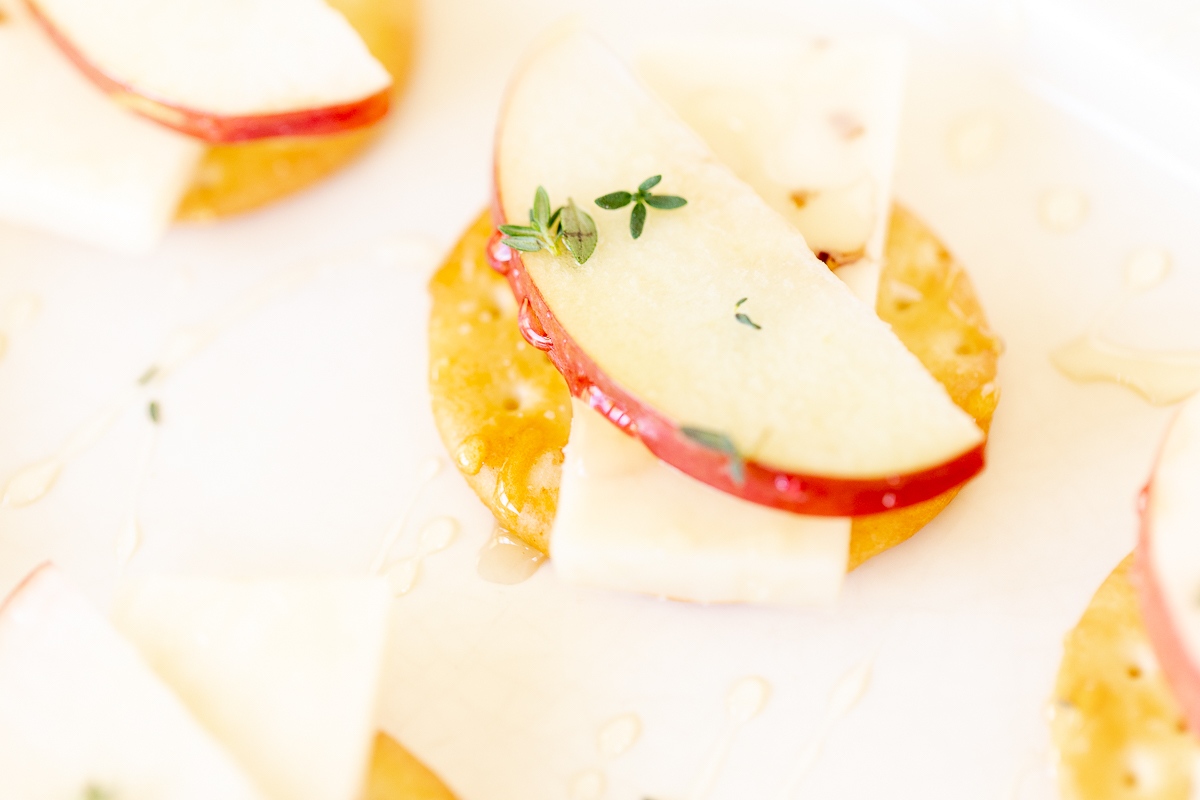 Cheese and crackers on a white platter, with an apple slice and fresh herbs.