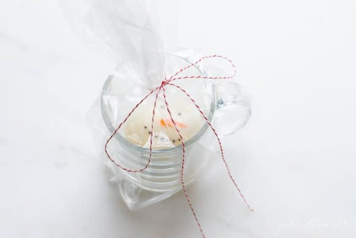A clear mug with a Christmas hot chocolate bomb inside, wrapped in cellophane and a baker's twine bow.
