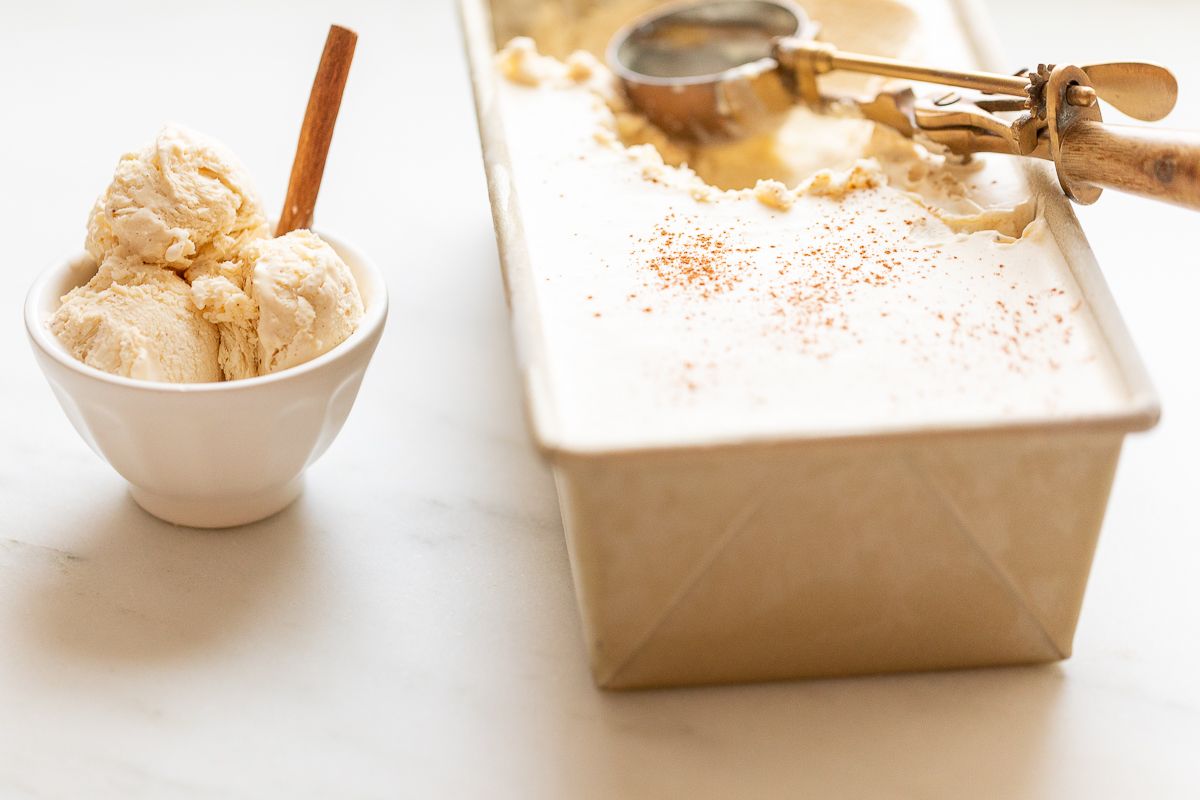 eggnog ice cream served in a small white bowl, with a gold loaf pan full of ice cream in the background.