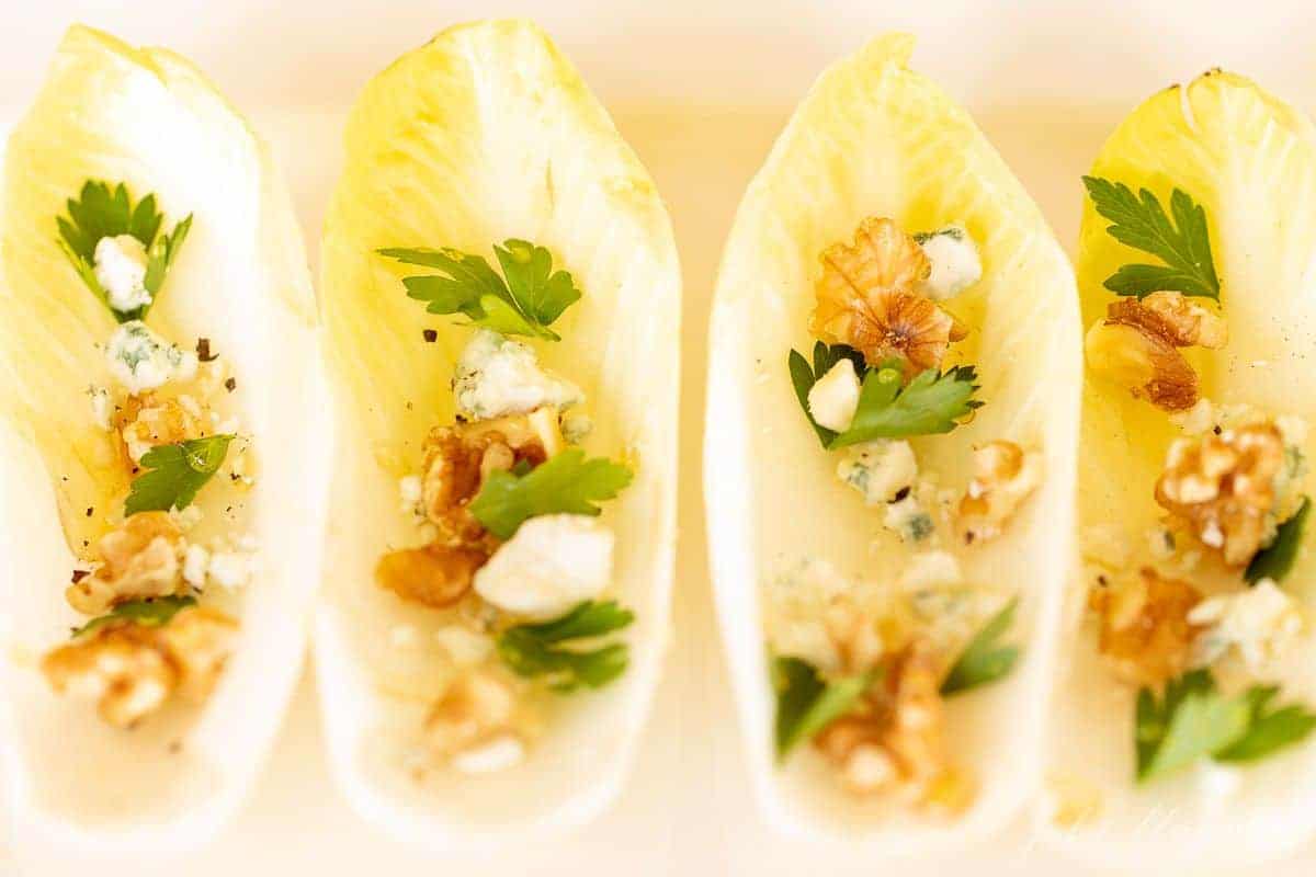 A white platter with four individual endive leafs, filled with toppings for a unique hors d'oeuvre.