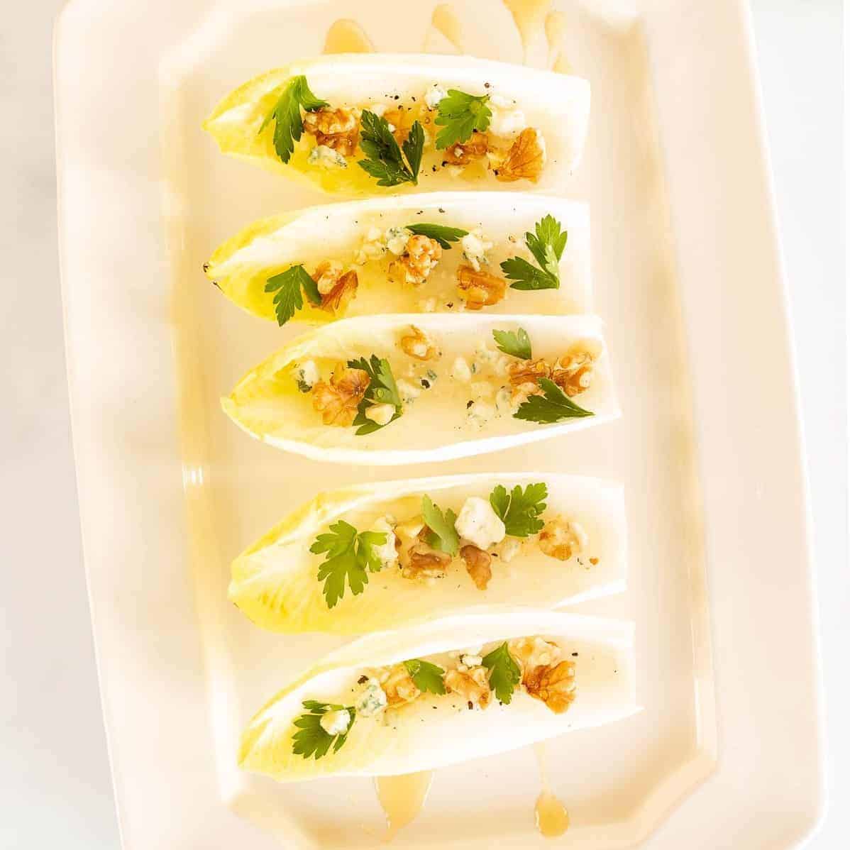 A white platter with individual endive lettuce leaves, filled with nuts, cheese and more for an endive salad appetizer.