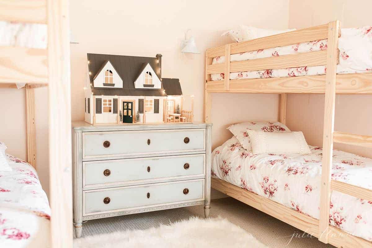 A little girls' room with bunk beds and a blue chest of drawers topped with a dollhouse decorated for Christmas.