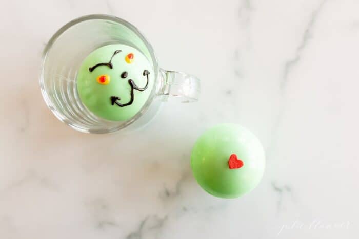 A green grinch hot chocolate bomb in a clear glass mug, a second bomb to the side on a marble surface.