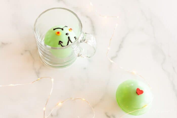 A green grinch hot chocolate bomb in a clear glass mug, a second bomb to the side on a marble surface.