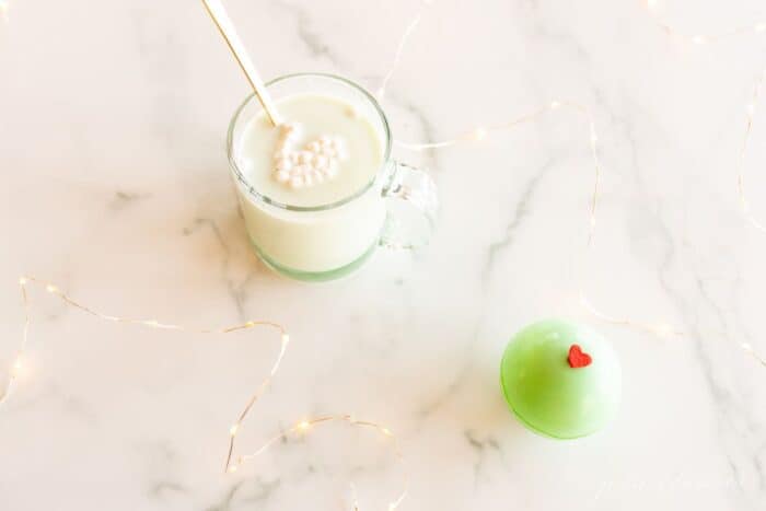 A clear glass mug of grinch hot chocolate with a green grinch hot chocolate bomb to the side.