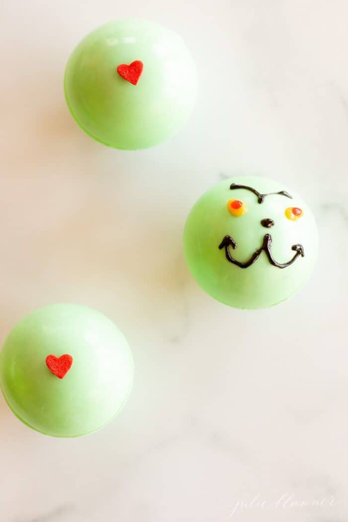 A green grinch hot chocolate bomb in a clear glass mug, more bombs to the side on a marble surface.