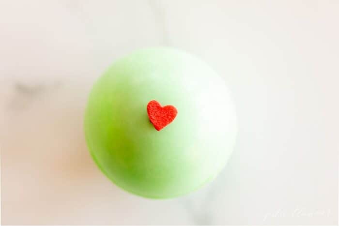 A single green grinch hot chocolate bomb decorated with a red heart, on a marble surface.