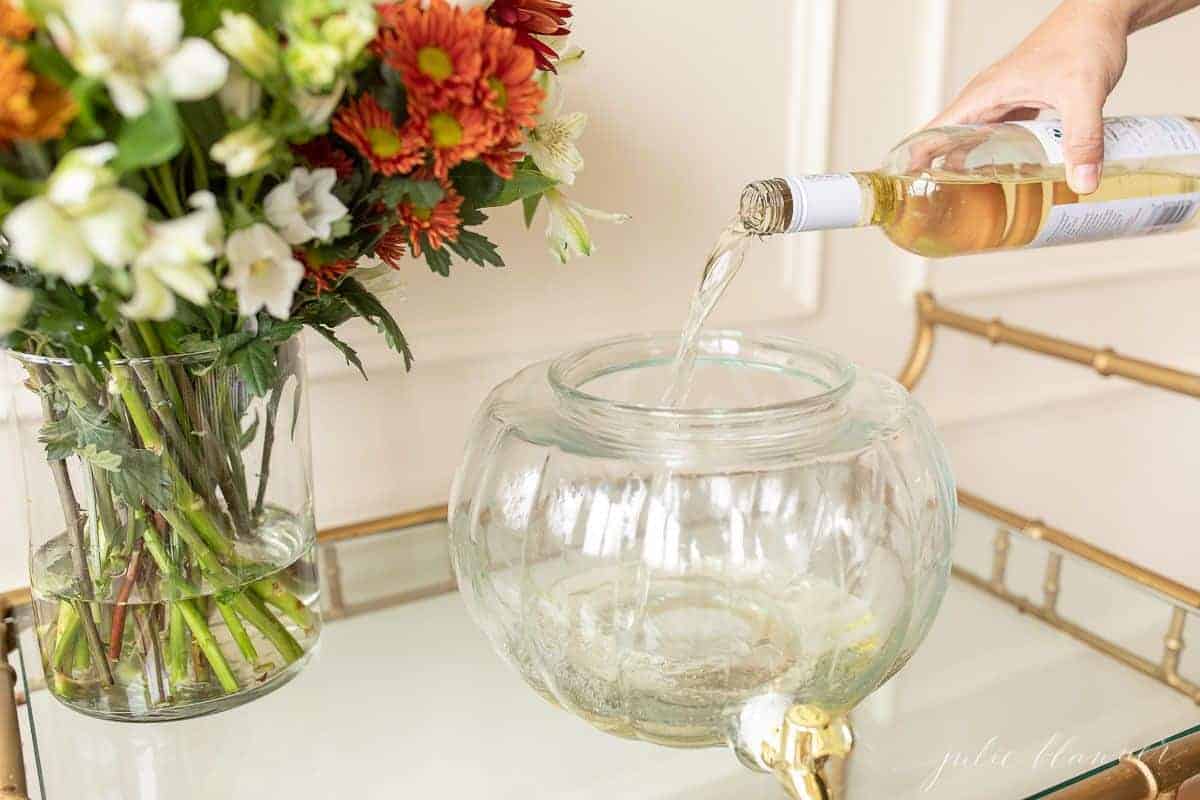 A glass bar cart holding a pumpkin drink dispenser, with a glass of wine being poured inside.