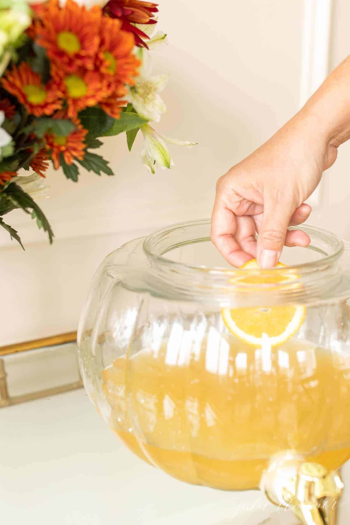 A glass bar cart holding a pumpkin drink dispenser, with a hand placing orange slices inside of a fall sangria mixture.