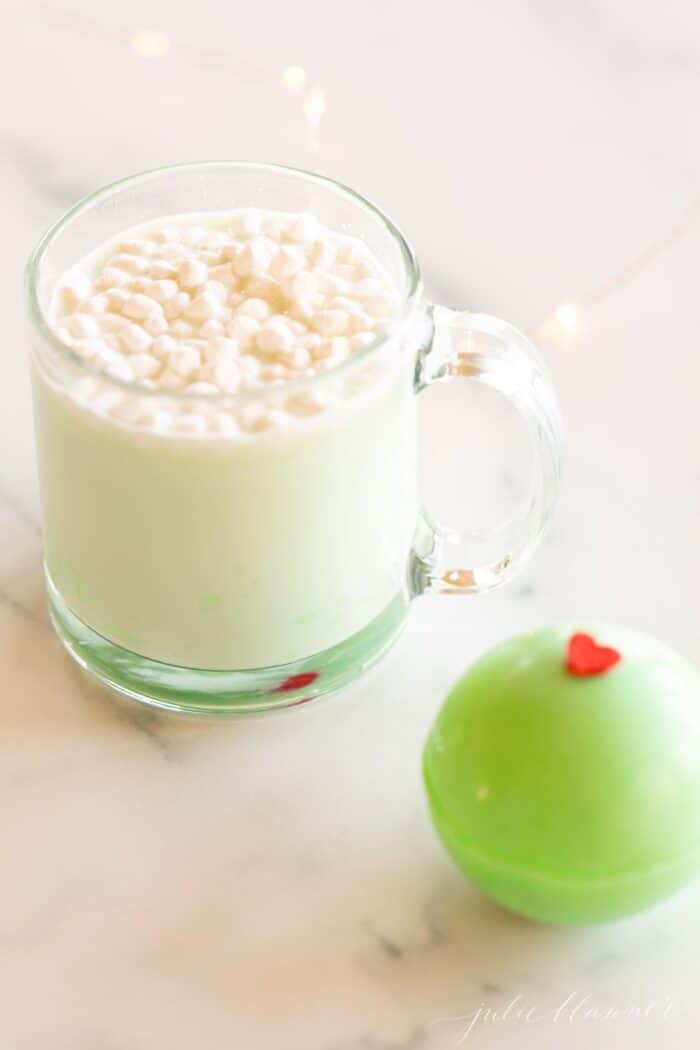 A clear glass mug of grinch hot chocolate with a green grinch hot chocolate bomb to the side.