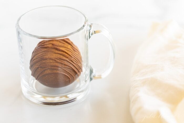 A clear mug with a spicy hot chocolate bomb inside, cinnamon stick and white linen napkin to the side.