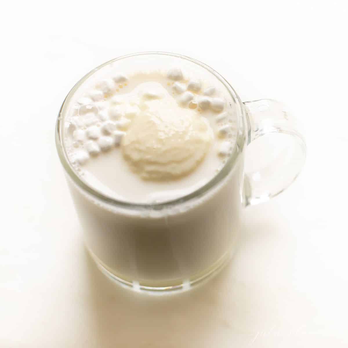 A clear mug of white hot chocolate with little marshmallows at the top.