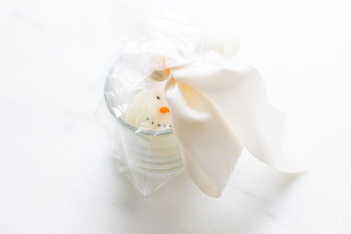 A clear mug with a Christmas hot chocolate bomb inside, wrapped in cellophane and a big white satin bow.