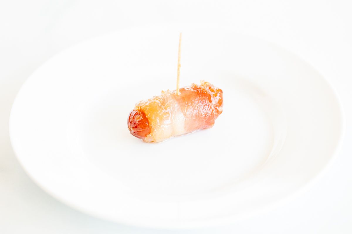 A single bacon wrapped little smokie on a white plate.