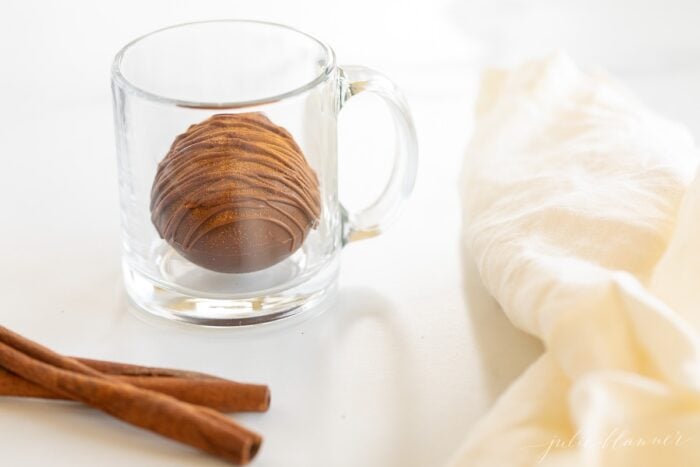 A clear mug with a Mexican hot chocolate bomb inside, cinnamon stick and white linen napkin to the side.