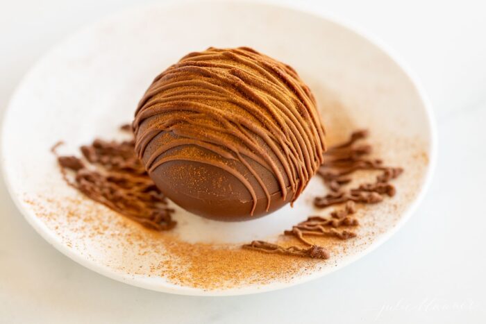 A white plate with a single Mexican hot chocolate bomb drizzled in cayenne pepper and chocolate.