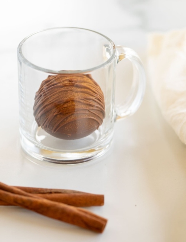 A Mexican hot chocolate bomb in a clear glass mug, with cinnamon sticks in the foreground.