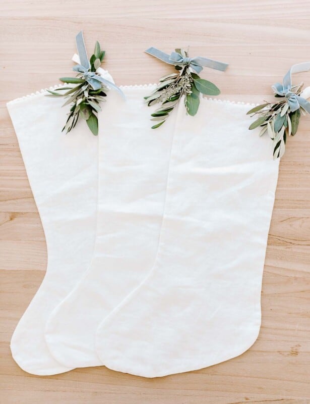 Three white linen stockings with touches of holiday greenery, laid out on a wooden surface.