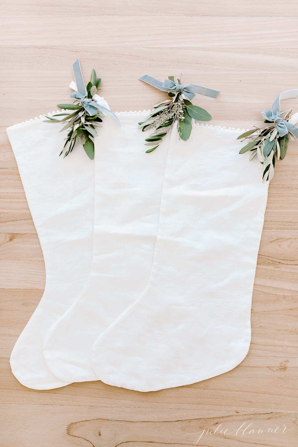 Three white linen stockings with touches of holiday greenery, laid out on a wooden surface.