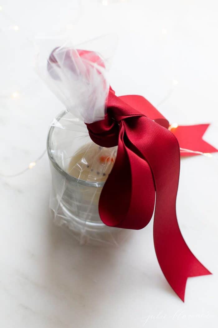 A clear mug with a Christmas hot chocolate bomb inside, wrapped in cellophane and a big red bow.