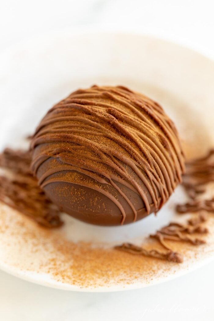A white plate with a single Mexican hot chocolate bomb drizzled in cayenne pepper and chocolate.
