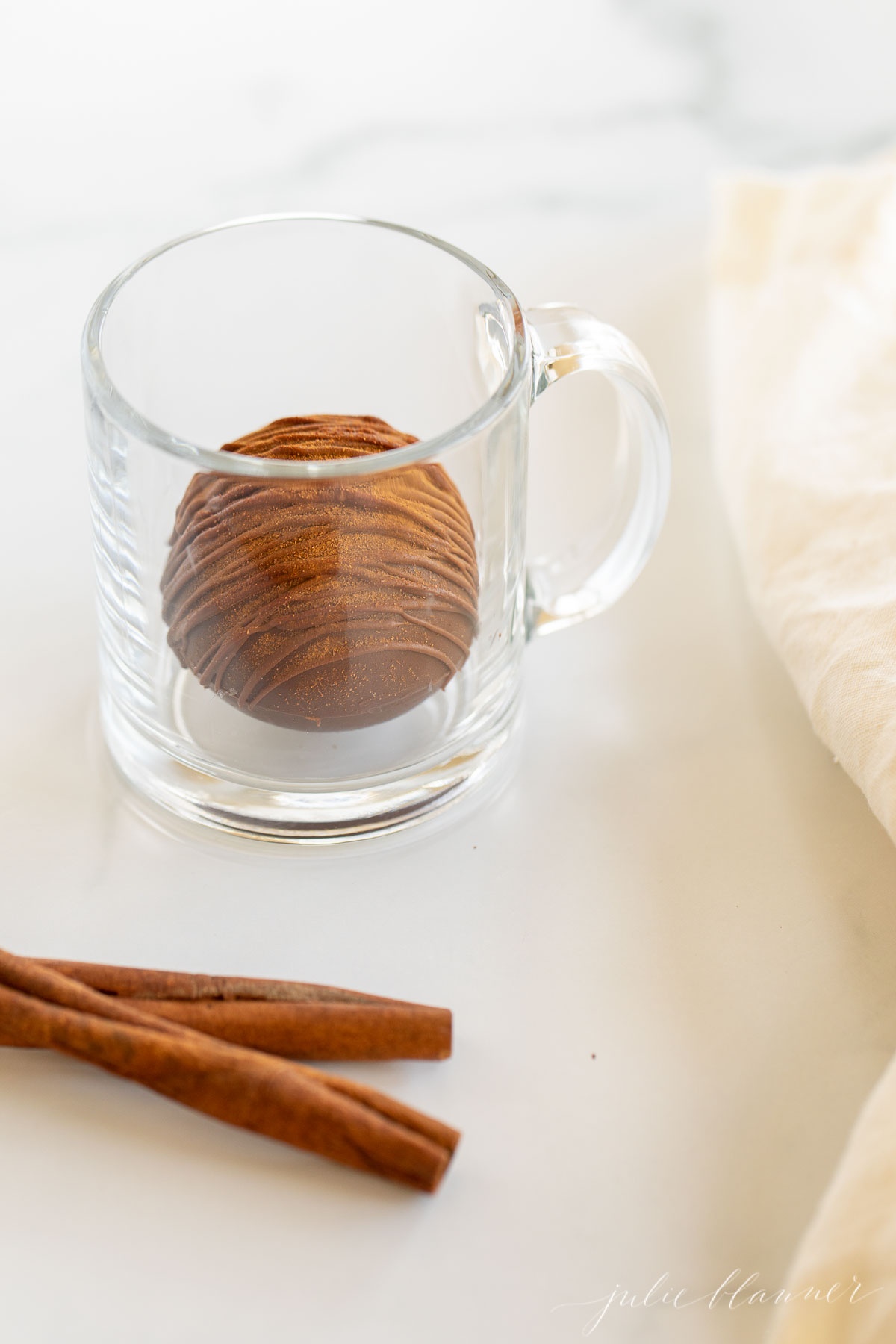 A clear mug with a Mexican hot chocolate bomb inside, cinnamon stick and white linen napkin to the side.