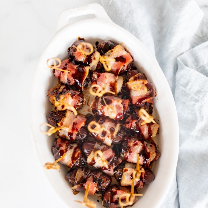 A white oval baking dish with bacon wrapped dates, each has a toothpick for serving.