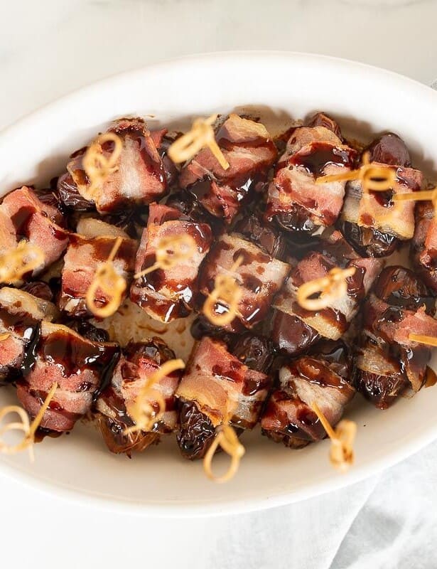 A white oval baking dish filled with dates wrapped in bacon, toothpicks in each one.