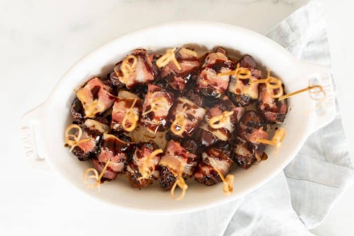 A white oval baking dish filled with dates wrapped in bacon, toothpicks in each one.