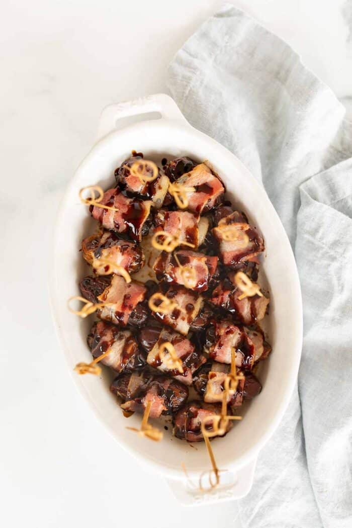 A white oval baking dish filled with dates wrapped in bacon hors d'oeuvres, toothpicks in each one.