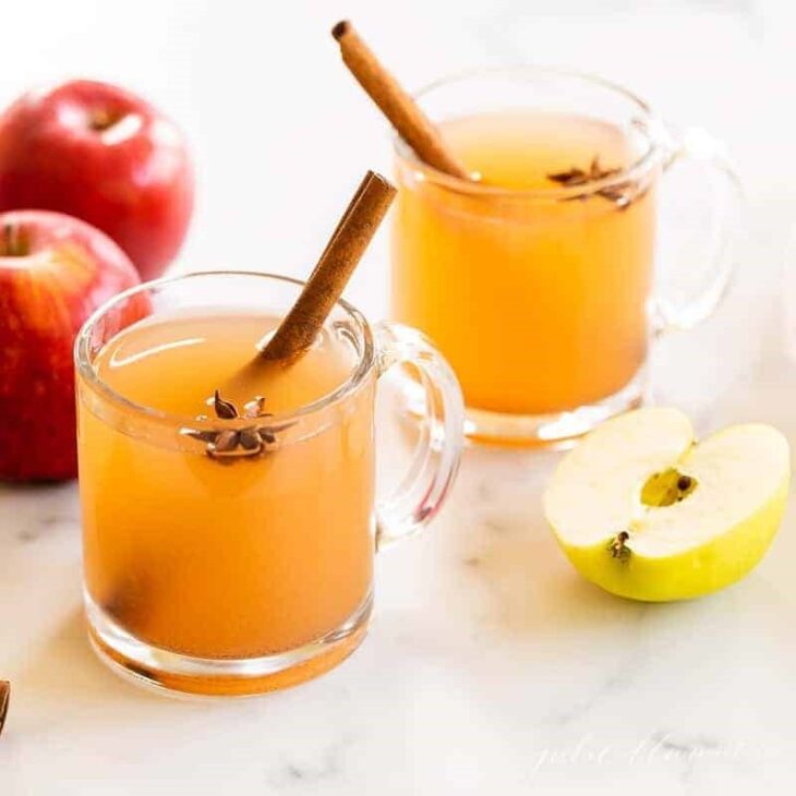 Two clear glass mugs full of Thanksgiving cocktails, cinnamon sticks and apples as garnish