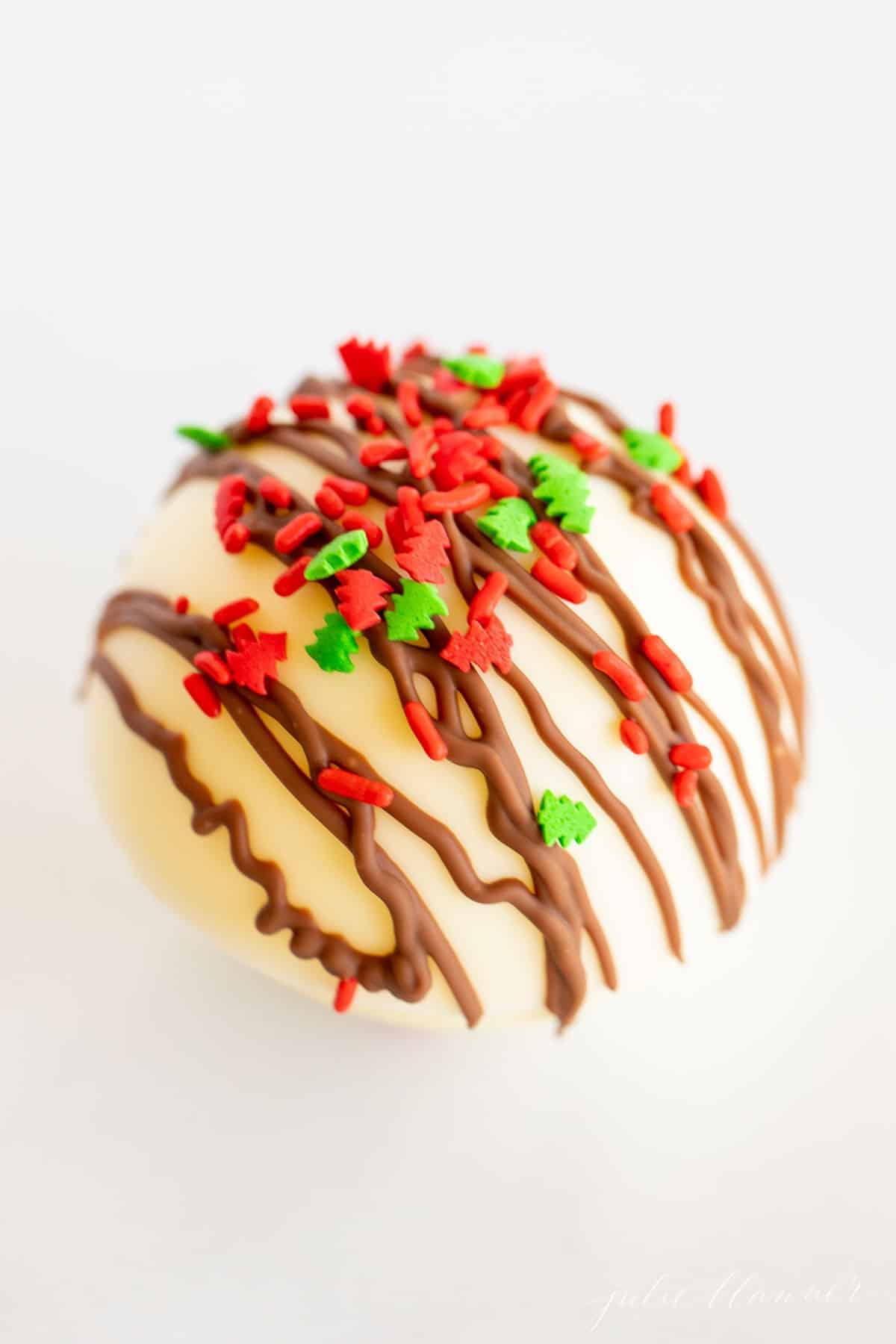 A white plate with a white chocolate hot chocolate bomb topped in Christmas sprinkles.