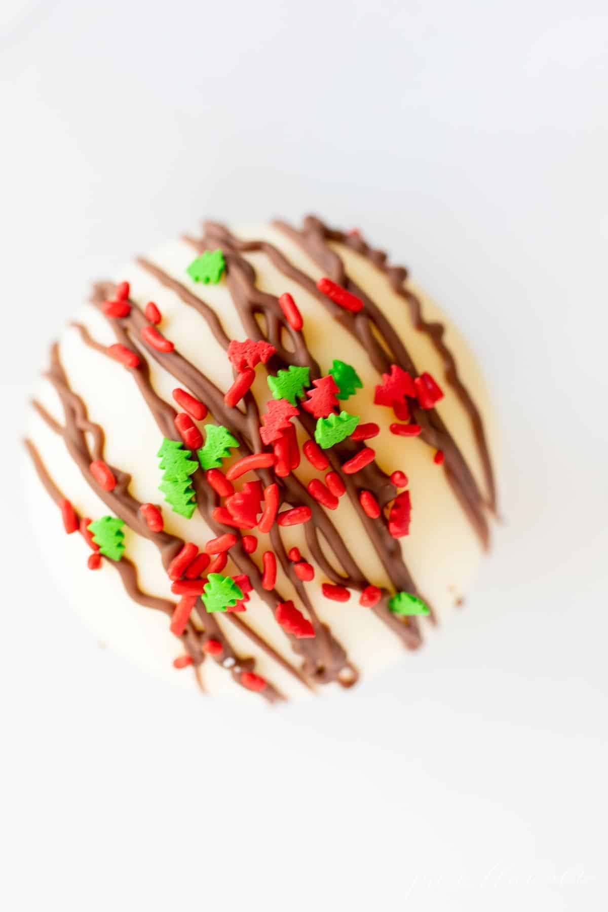 A white plate with a white chocolate hot chocolate bomb topped in Christmas sprinkles.