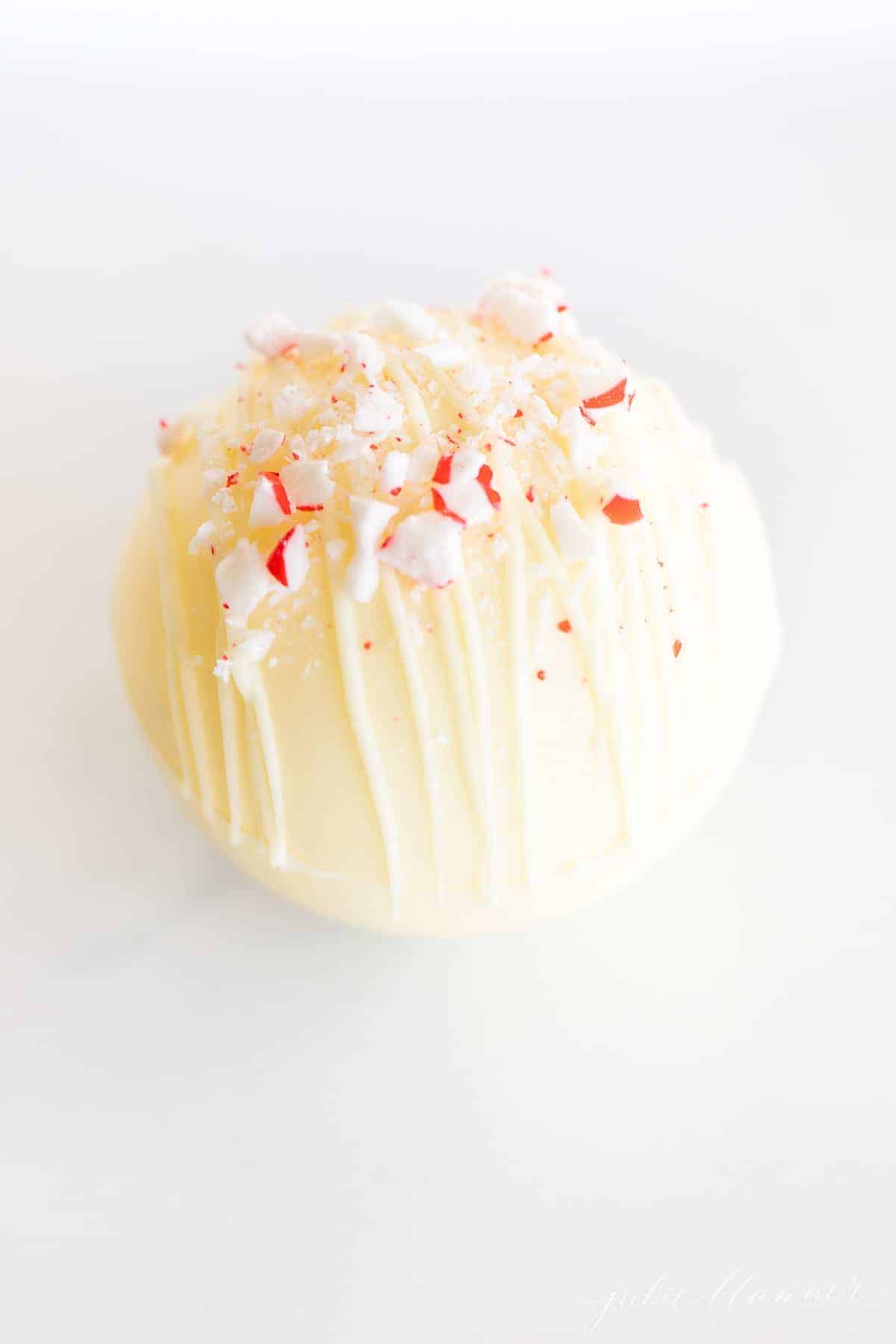 A white chocolate hot chocolate bomb covered in crushed peppermint on a marble surface.