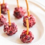 Mini cheese ball bites covered in dried cranberries, with a pretzel stick in the center, resting on a white platter.