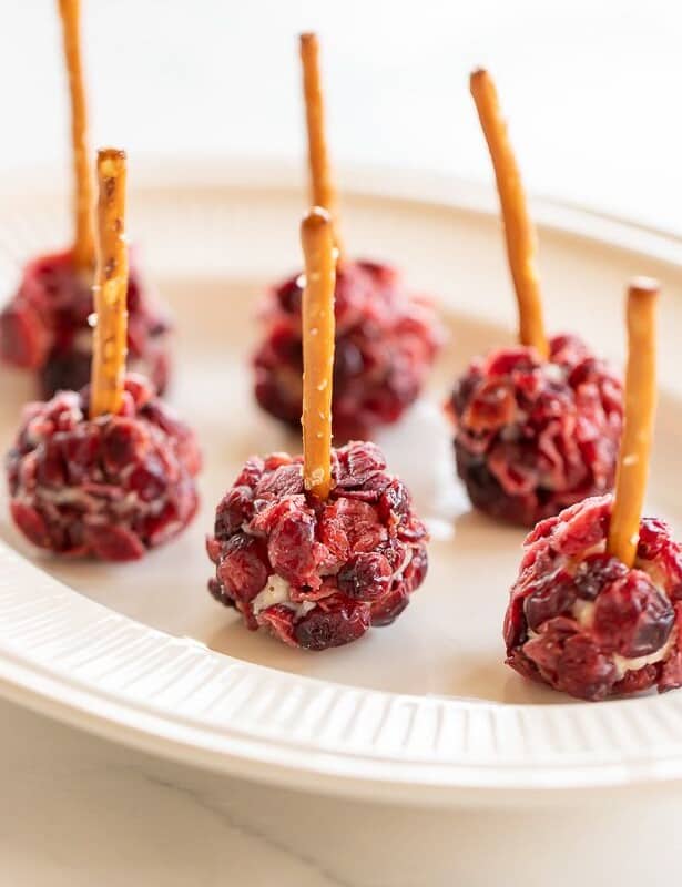Individual cheese ball bites, covered in dried cranberries with pretzel sticks sticking out of each on a white plate.