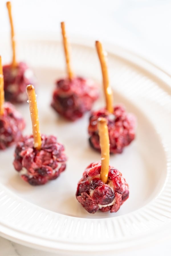 Mini cheese ball bites covered in dried cranberries, with a pretzel stick in the center, resting on a white platter.