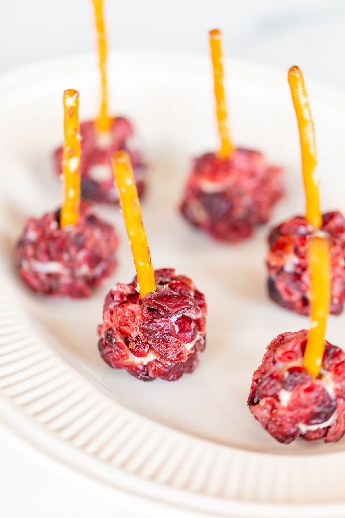Mini cheese ball bites covered in dried cranberries, with a pretzel stick in the center, resting on a white platter.
