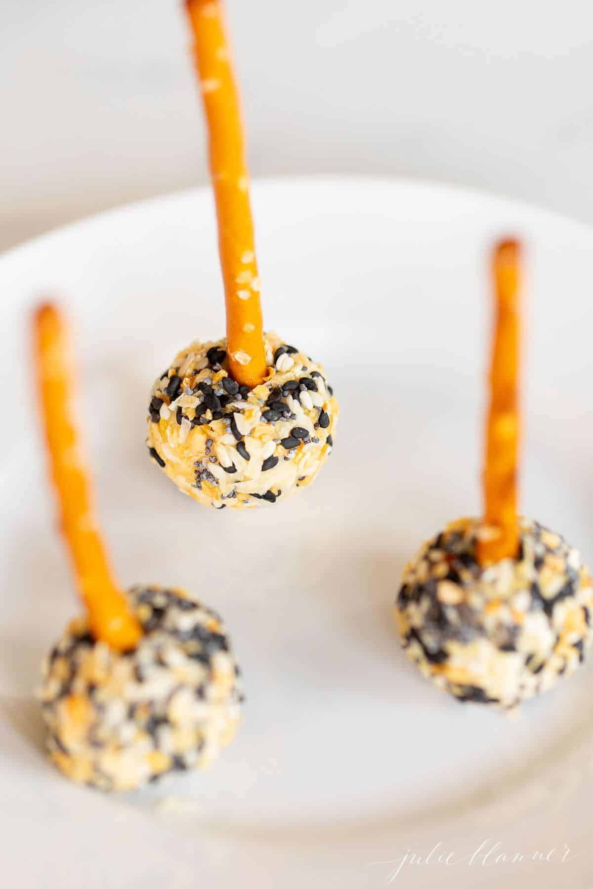 Mini cheese ball bites on a white plate, covered in everything bagel seasoning, pretzel sticks sticking out of each for serving.