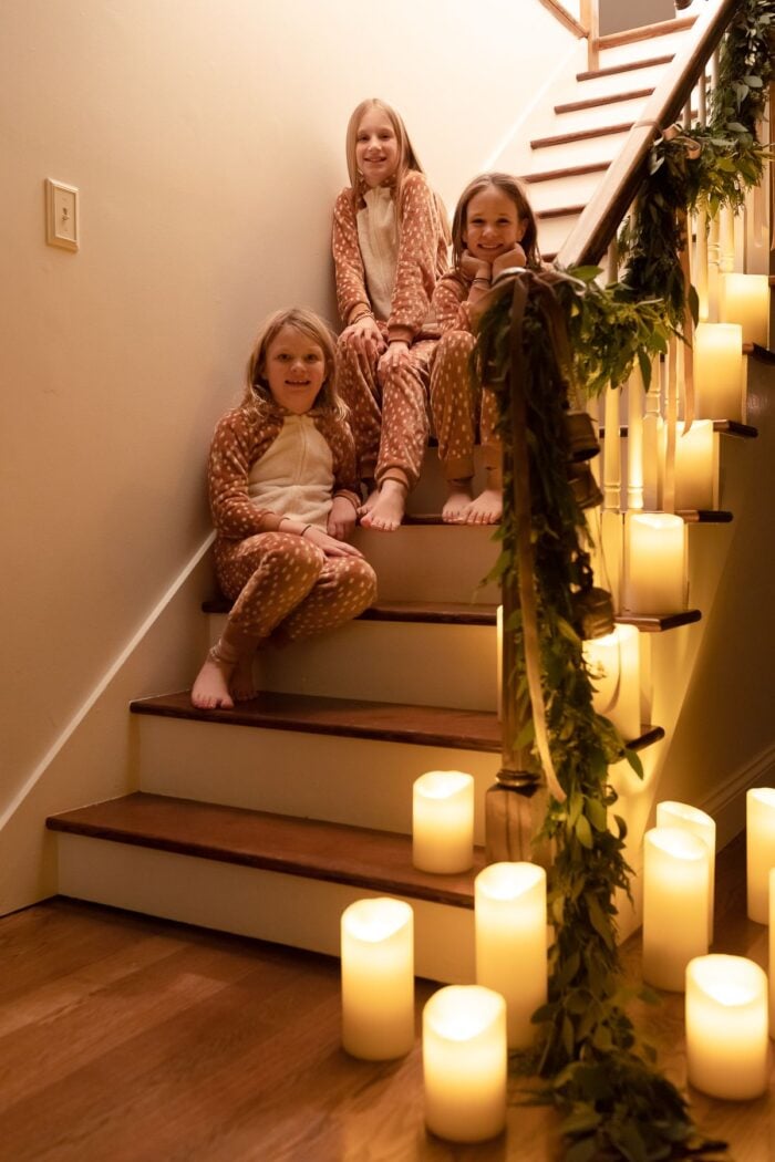 A garland on a staircase with pillar candles all around, three little girls on the stairs in Christmas pajamas