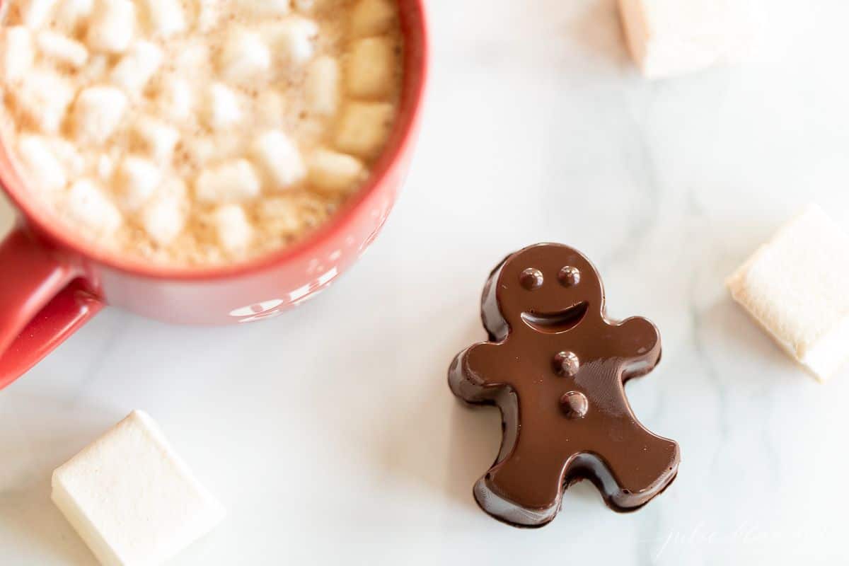 A red mug full of dark hot chocolate with marshmallows, with a dark chocolate gingerbread man to the side.