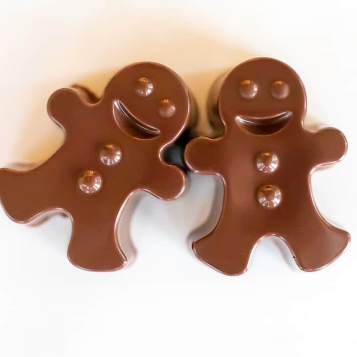 A pair of dark chocolate hot chocolate bombs shaped as gingerbread men on a white marble surface.