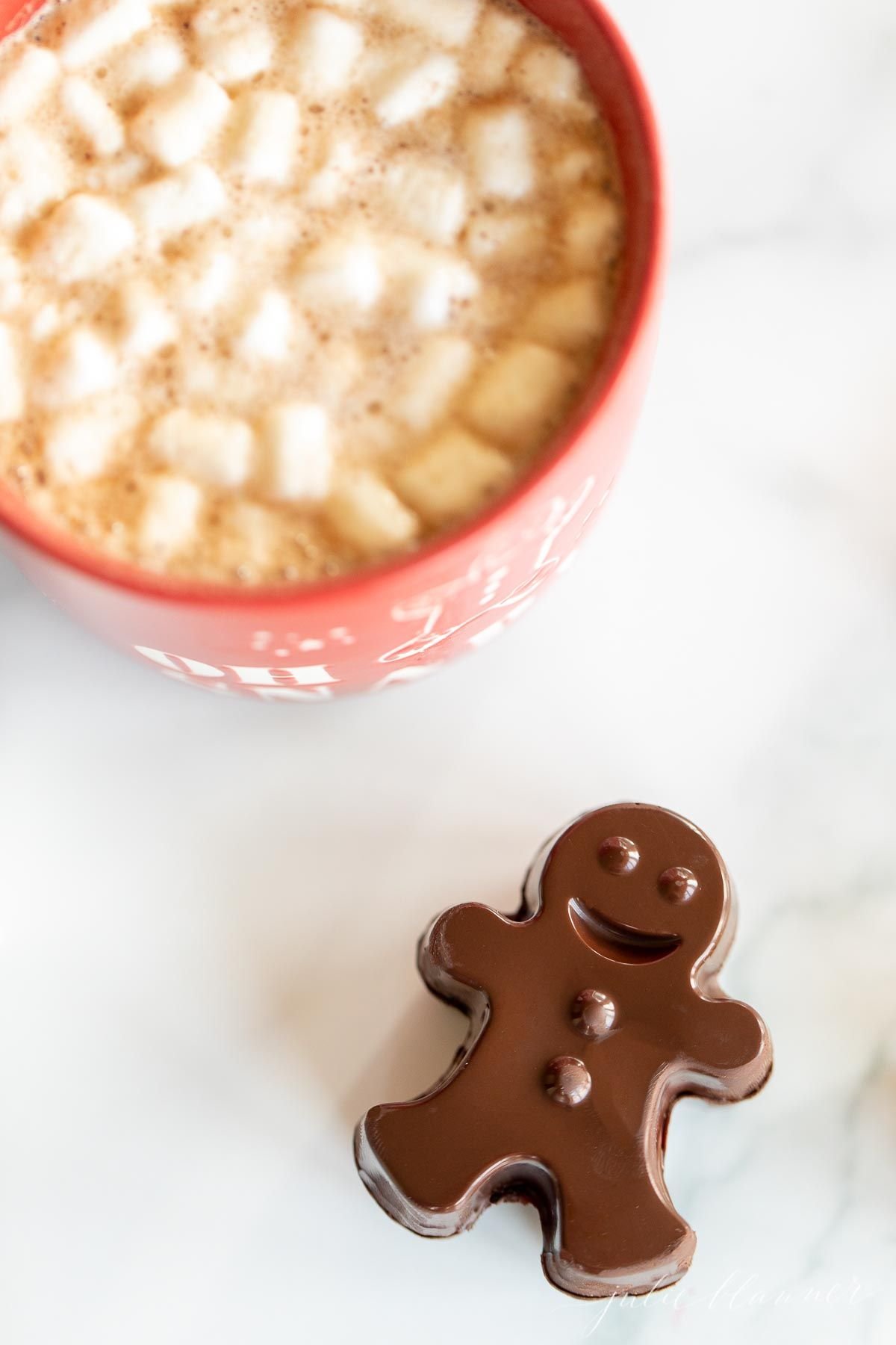 A red mug full of dark hot chocolate with marshmallows, with a dark chocolate gingerbread man to the side.