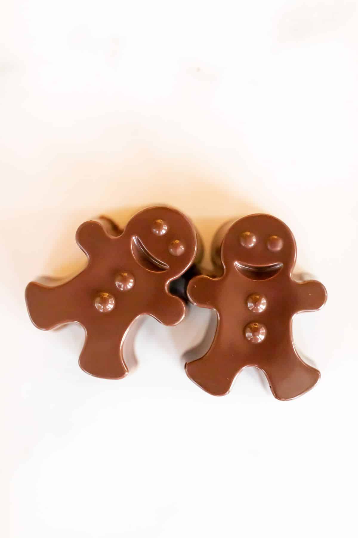 A pair of dark chocolate hot chocolate bombs shaped as gingerbread men on a white marble surface.